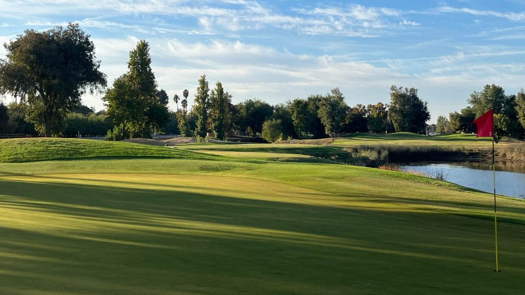 Red flag on a Lemoore Golf Course green beside water with fairway and trees in the background.