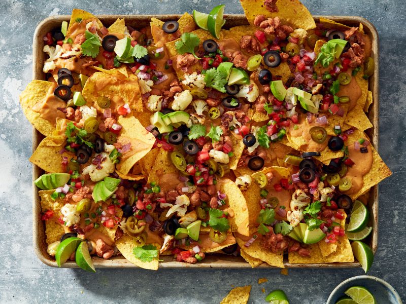 Loaded nachos topped with avocado, jalapeños, olives, pico de gallo, beans, cilantro, and lime wedges.