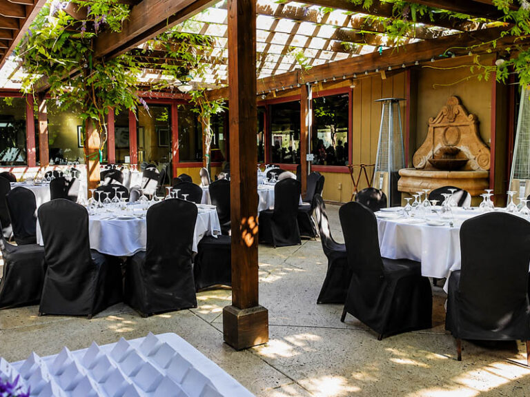 Outdoor wedding reception patio with round tables, white linens, black chair covers, and pergola greenery.